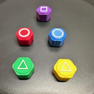 Könnte beinhalten: Fünf sechseckige Knöpfe mit verschiedenen farbigen Basen und weißen Symbolen. Die Knöpfe sind lila, rot, blau, grün und gelb. Die Symbole sind ein Quadrat, ein Kreis, ein Kreis mit einer Linie durch ihn, ein Dreieck und ein Dreieck mit einer Linie durch ihn.