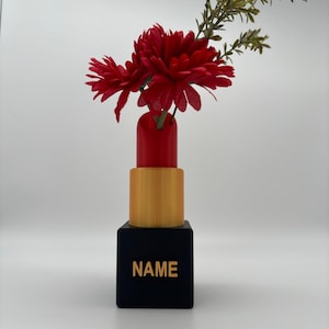 May include: A black lipstick tube vase with a gold band and the word "NAME" on the base. The vase is filled with red flowers and green foliage.