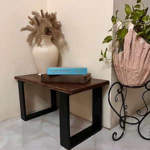 Puede incluir: Una pequeña mesa rectangular de madera con tapa marrón oscuro y patas negras. Un jarrón blanco con flores secas y una caja azul con el texto "COCONUT EXPERIENCE" están sobre la mesa. A la derecha hay un soporte decorativo para plantas con una maceta rosa.