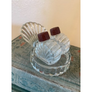 May include: A set of two clear glass salt and pepper shakers shaped like seashells with maroon tops. The shakers are sitting on a clear glass tray shaped like a seashell.