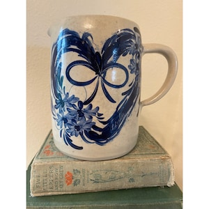 May include: A cream-colored ceramic pitcher with a blue and white floral design. The pitcher has a heart-shaped design with a blue bow in the center. The pitcher has a handle and a spout.