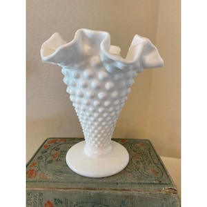 May include: White milk glass vase with a ruffled top and a textured, dotted body. The vase is sitting on a vintage book.