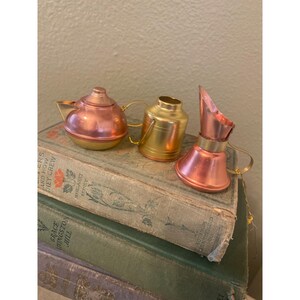 May include: Three miniature metal pitchers in gold and copper tones. The pitchers are arranged on a stack of vintage books.