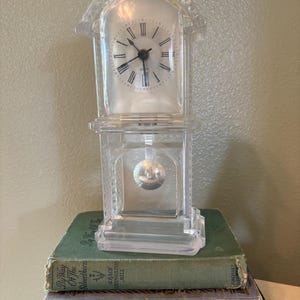 May include: A clear, plastic clock with a rectangular base and a domed top, featuring a clock face with black Roman numerals. The clock is perched atop a stack of vintage books with green and gray covers.