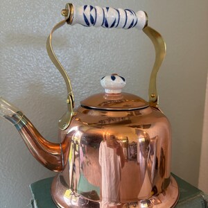 May include: A copper tea kettle with a brass handle and spout. The handle is white with blue floral designs. The lid has a matching white and blue knob. The kettle is sitting on top of a stack of books.