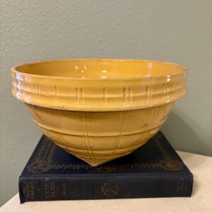 May include: A yellow ceramic mixing bowl with a textured, grid-like pattern. The bowl is set on a dark blue book with gold lettering. The bowl has a wide rim and a rounded shape, perfect for kitchen use.