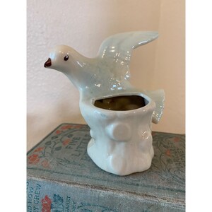 May include: A white ceramic bird planter with a blue glaze. The bird is perched on a tree stump with a small hole for planting.