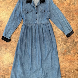 May include: A light blue denim dress with long sleeves, a black velvet collar, and black cuffs. The dress features two chest pockets with button closures and a button-down front. The skirt is gathered at the waist.