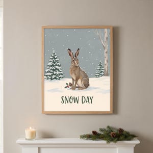 May include: A framed wall art print featuring a brown hare sitting in a snowy landscape with evergreen trees, a birch tree, and holly berries. The scene is set against a grey sky with falling snow, and the text SNOW DAY is printed in green.