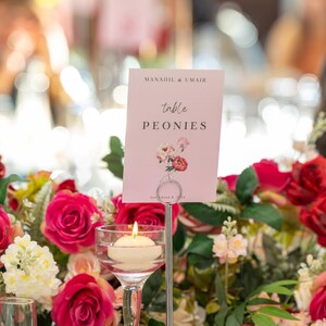 May include: A pink table number card with the text "Manadie & Umair" and "table PEONIES" in black font. The card is decorated with a floral illustration of pink and white peonies. The card is placed on a silver stand in front of a floral arrangement of red and white roses.
