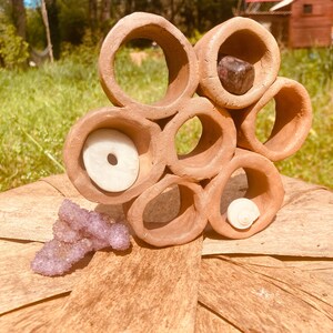 May include: A terracotta clay shelf with eight circular openings, each containing a different natural object, including a white shell, a purple stone, and a white disc with a hole in the center. The shelf is sitting on a wooden surface.