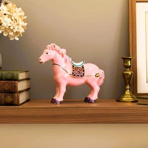 May include: A pink ceramic horse figurine featuring a decorative saddle with white, teal, and dark red accents, gold-toned trim, and dark purple hooves. The horse stands in a profile position, showcasing a smooth finish and detailed mane and tail.