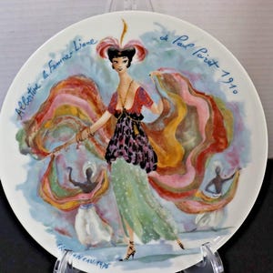 May include: A decorative plate featuring a colorful illustration of a woman in a flowing gown, with text that reads "Albertine la Femme Liane" and "de Paul Poiret 1910". The plate has a white background and is held by a clear stand.