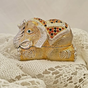 May include: A decorative elephant figurine in gold and silver tones, adorned with orange, black, and clear gemstones. The elephant has a green gemstone eye and is resting on a white lace surface. The figurine is a jewelry box.
