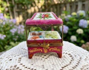 May include: A rectangular porcelain trinket box with a magenta base and lid, featuring gold-tone metal trim and a latch. The lid is decorated with a floral motif, while the interior base displays painted illustrations of carrots and green vegetables.