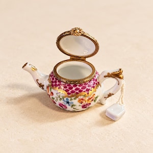 May include: A miniature porcelain teapot trinket box with a hinged lid, featuring a white base with a pink scale pattern, gold trim, and colorful floral motifs. A small white ceramic tea bag tag hangs from the handle by a thin white string.