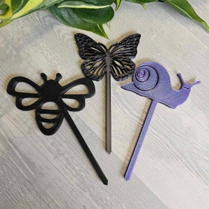 May include: Three garden markers made of black and purple plastic. The markers are shaped like a bee, a butterfly, and a snail.