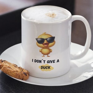 Puede incluir: Taza de cerámica blanca con un pato de dibujos animados con gafas de sol y el texto "I DON'T GIVE A DUCK". La taza está llena de una bebida espumosa y se encuentra sobre un platillo blanco. Una galleta con chispas de chocolate descansa en el platillo.