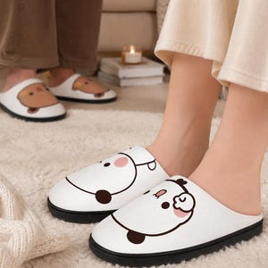 May include: White house slippers with a black sole feature a cartoon panda face design. The slippers are worn by a person standing on a white rug. Another person wears similar slippers in the background.