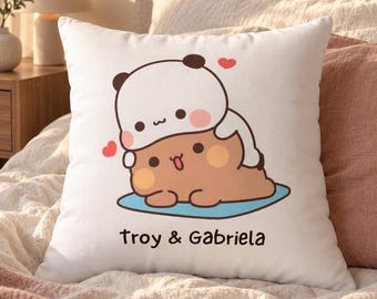 May include: A square white throw pillow featuring a cartoon illustration of a white panda sitting on top of a brown bear. The bears are on a blue oval shape with two small red hearts above them. The text Troy & Gabriela is printed in black below.