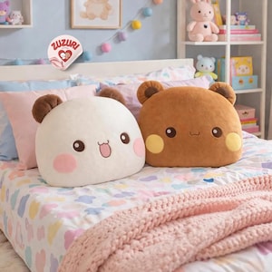 May include: Two plush bear-shaped pillows with smiling faces rest on a bed with a pastel floral patterned comforter. One pillow is white with pink cheeks, the other is brown with yellow cheeks. The room is decorated with stuffed animals and a Zuzuri sticker.