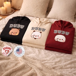 Puede incluir: Tres sudaderas con capucha en negro, crema y burdeos, cada una con la palabra "BUBU" encima de una cara de oso de dibujos animados. Las sudaderas con capucha están dobladas cuidadosamente sobre una cama con almohadas y una manta. La imagen también incluye dos pegatinas redondas.