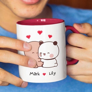 Puede incluir: Taza de cerámica blanca con borde y asa rojos. La taza presenta una ilustración de dibujos animados de dos osos abrazándose con corazones por encima. El texto "Mark ♥ Lily" está impreso debajo de los osos.