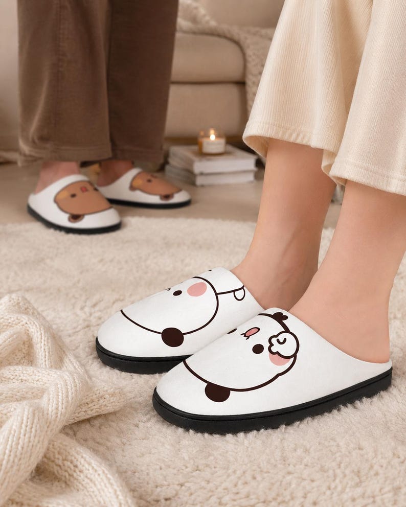 May include: White house slippers with a black sole feature a cartoon panda face design. The slippers are worn by a person standing on a white rug. Another person wears similar slippers in the background.