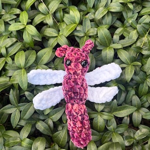 May include: A handmade crochet dragonfly plush toy featuring a textured, variegated dark pink and purple body with two pairs of soft white wings. The insect has two small, round, black plastic eyes and two small antennae on its head.