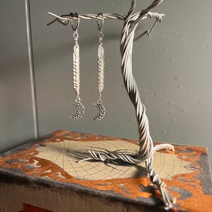 May include: A pair of silver earrings with clear quartz crystals wrapped in silver wire and a crescent moon charm hanging from each earring. The earrings are displayed on a silver wire tree.