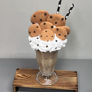 May include: A chocolate milkshake in a clear glass, topped with whipped cream and chocolate chip cookies. Two black and white polka dot straws are in the milkshake. The milkshake sits on a small, stained wooden tray.