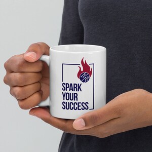 May include: White ceramic mug with the text "SPARK YOUR SUCCESS" in navy blue, along with a graphic of a circuit board inside a flame. The mug is held by a person.