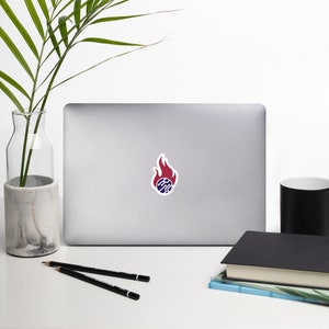May include: A laptop computer with a sticker of a red flame with a blue circle inside. The circle has a pattern of lines inside.