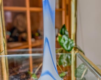 May include: A tall, slender glass vase with a white base and a blue, swirling design. The vase has a clear, rounded base and a long, cylindrical neck. The blue design flows from the base to the top, creating a decorative pattern. The vase is approximately 12 inches tall.