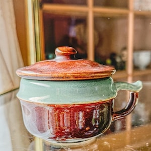 May include: A small brown and green ceramic pot with a lid and a handle. The pot has a glazed finish and a rustic look.