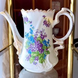 May include: A white porcelain teapot with a gold trim and a floral design featuring purple and blue flowers. The teapot has a curved handle and a spout.