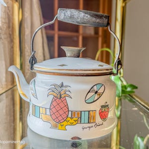 May include: White enamel teapot with a metal handle and spout. The teapot is decorated with a pineapple, strawberry, and geometric designs. The handle is made of metal and has a wooden grip. The teapot has the text "Georges Briard" on the side.