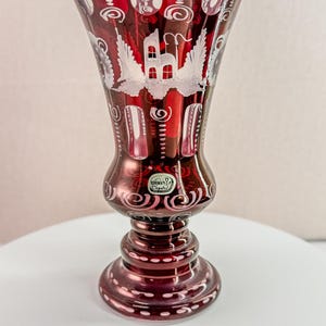 May include: A ruby red Bohemian crystal vase with a flared top and a detailed base. The vase features white etched designs of a building with wings and decorative swirls. The vase is approximately 8 inches tall.