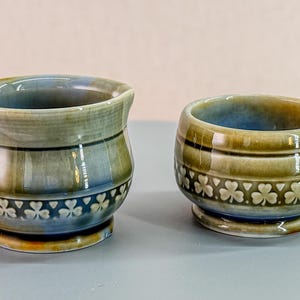 May include: Two ceramic pieces, a small pitcher with a handle and a small cup, both in shades of blue, green, and brown. Each piece features a band of shamrock designs around the base. The pitcher has a handle and a spout.