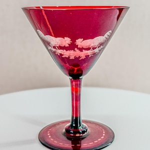 May include: A ruby red cocktail glass with a triangular bowl and a long stem. The glass features an etched design of two roosters facing each other. The base of the glass is round with a dotted design. A vintage-style glass.
