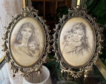 May include: Two gold-toned oval picture frames with ornate details. Each frame holds a black and white drawing of a woman and child. The drawing in the left frame shows a woman with long hair holding a child. The drawing in the right frame shows a woman with long hair holding a child in her arms.