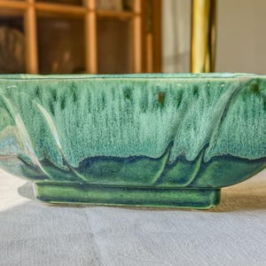 May include: An elongated ceramic bowl featuring a glossy glaze in shades of deep forest green and lighter mint green. The vessel has a rectangular base and curved, scalloped sides with a vertical drip pattern throughout the exterior surface.