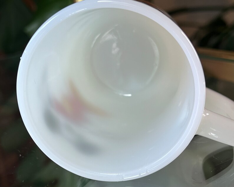 以下が含まれることがあります： 上から見た、白いセラミックマグカップ。マグカップの内側は白く、底にはかすかなデザインが見えます。マグカップはぼやけた背景に置かれています。