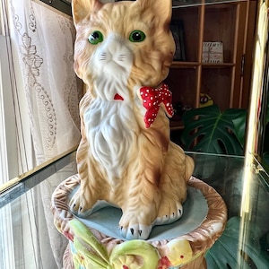 May include: A ceramic figurine of a cat wearing a red bow with white polka dots. The cat is sitting on a base decorated with flowers and a green bow. The figurine is a vintage style.