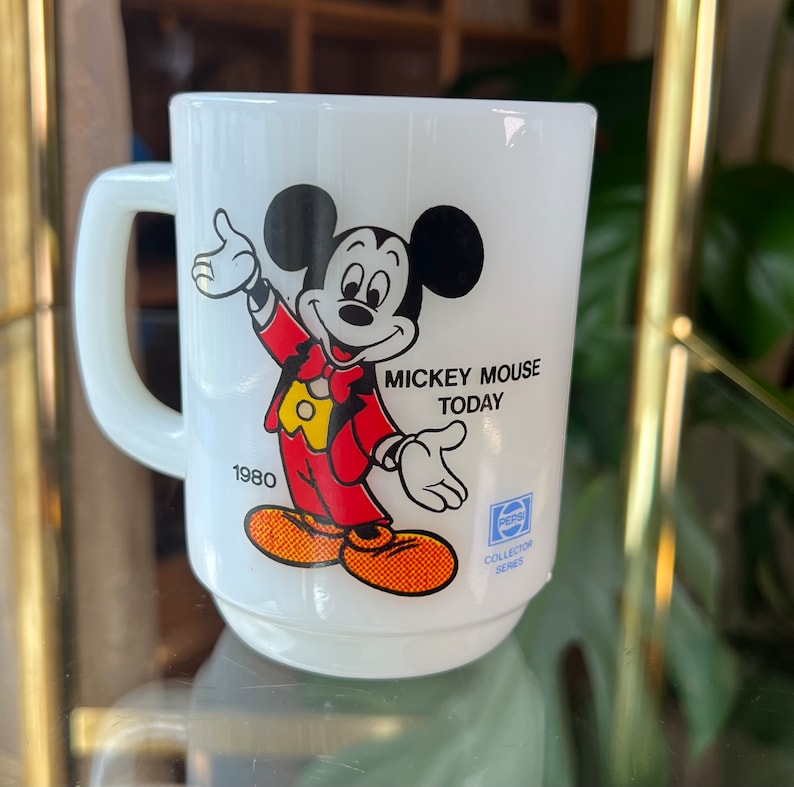 以下が含まれることがあります： 赤色のジャケットと黄色のシャツを着たミッキーマウスが描かれた白いセラミックマグ。マグには「Mickey Mouse Today」と「1980」の文字が入っています。ペプシコレクターシリーズのロゴも表示されています。マグには取っ手が付いています。