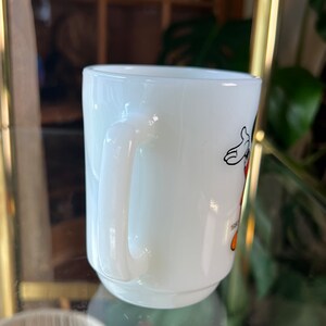 以下が含まれることがあります： 白いセラミックマグカップ、湾曲したハンドル付き。 マグカップには、黒、赤、黄色の漫画のキャラクターイラストが描かれています。 マグカップはぼやけた背景に配置されています。