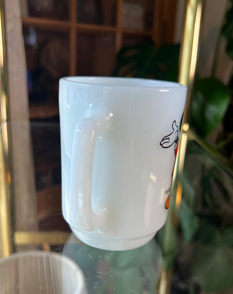 以下が含まれることがあります： 白いセラミックマグカップ、湾曲したハンドル付き。 マグカップには、黒、赤、黄色の漫画のキャラクターイラストが描かれています。 マグカップはぼやけた背景に配置されています。