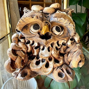 May include: A detailed, handcrafted owl sculpture in shades of brown and tan. The owl has large, black eyes with gold-colored rings, a yellow beak, and textured, layered feathers. The sculpture is made of a ceramic-like material.