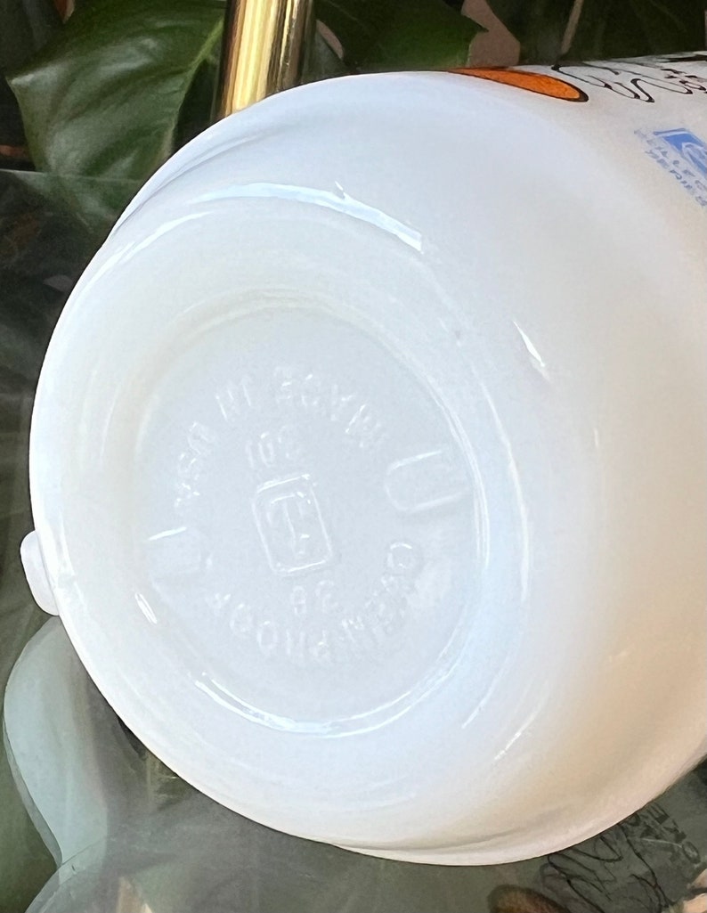 以下が含まれることがあります： 丸みを帯びた底の白いセラミックマグ。マグの底には「MADE IN USA」と「OVENPROOF 36」のエンボス加工された文字があります。マグは滑らかで光沢のある仕上がりです。