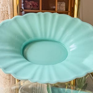 May include: A light blue, oval-shaped serving dish with a scalloped edge. The dish has a fluted design radiating from the center. The dish is made of a solid, opaque material.